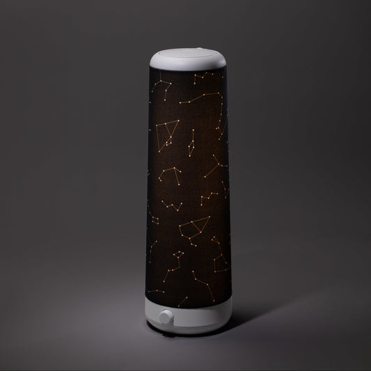 A cylindrical Nightside lamp with a Starlight Cotton Blackout Shade, featuring golden constellation patterns, stands on a plain gray surface against a gray background.