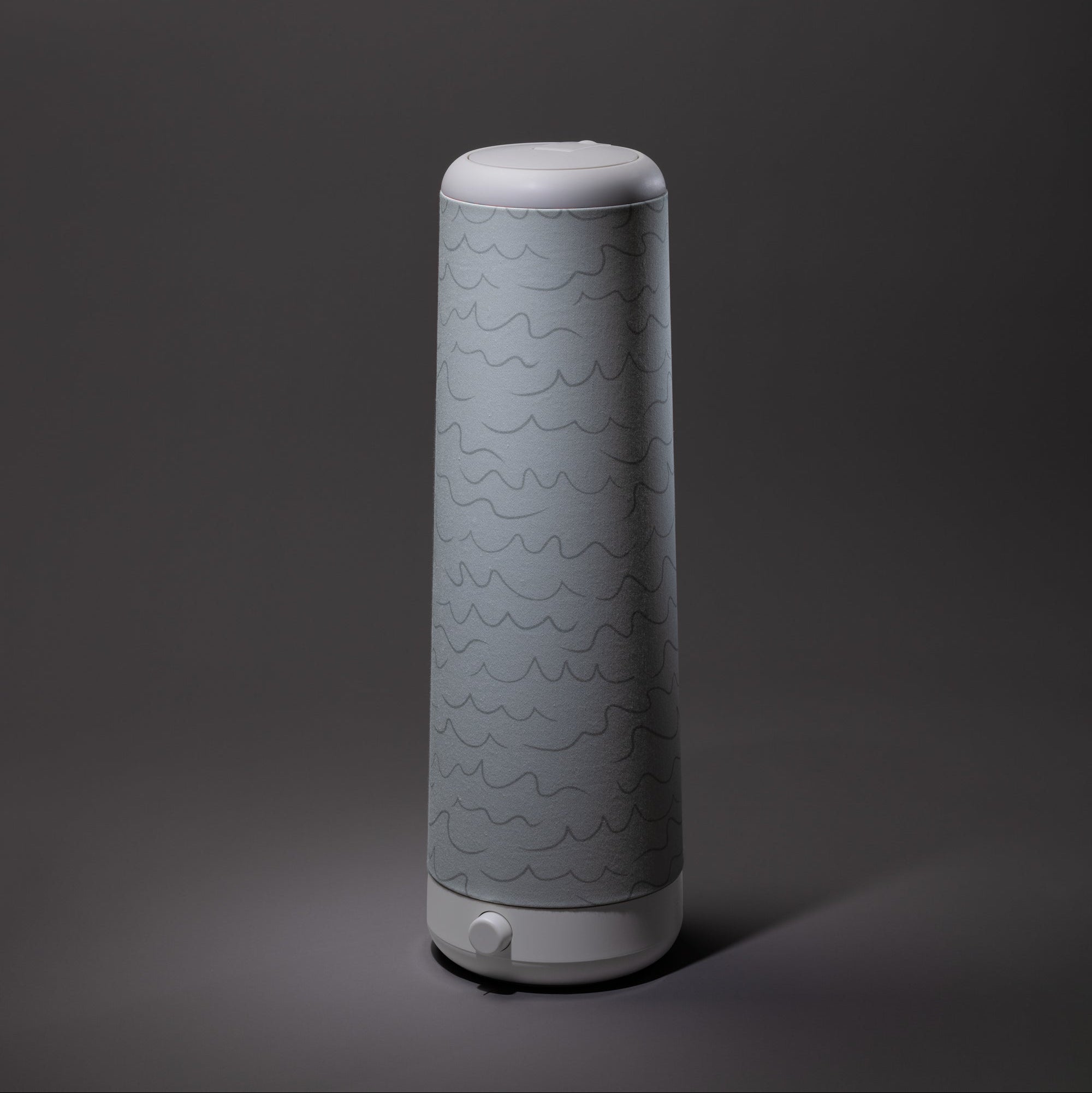A sleek, cylindrical light grey Cursive Lines Cotton Shade by Nightside with subtle wavy patterns stands on a dark grey background, resembling a modern lamp with interchangeable fabric shades.