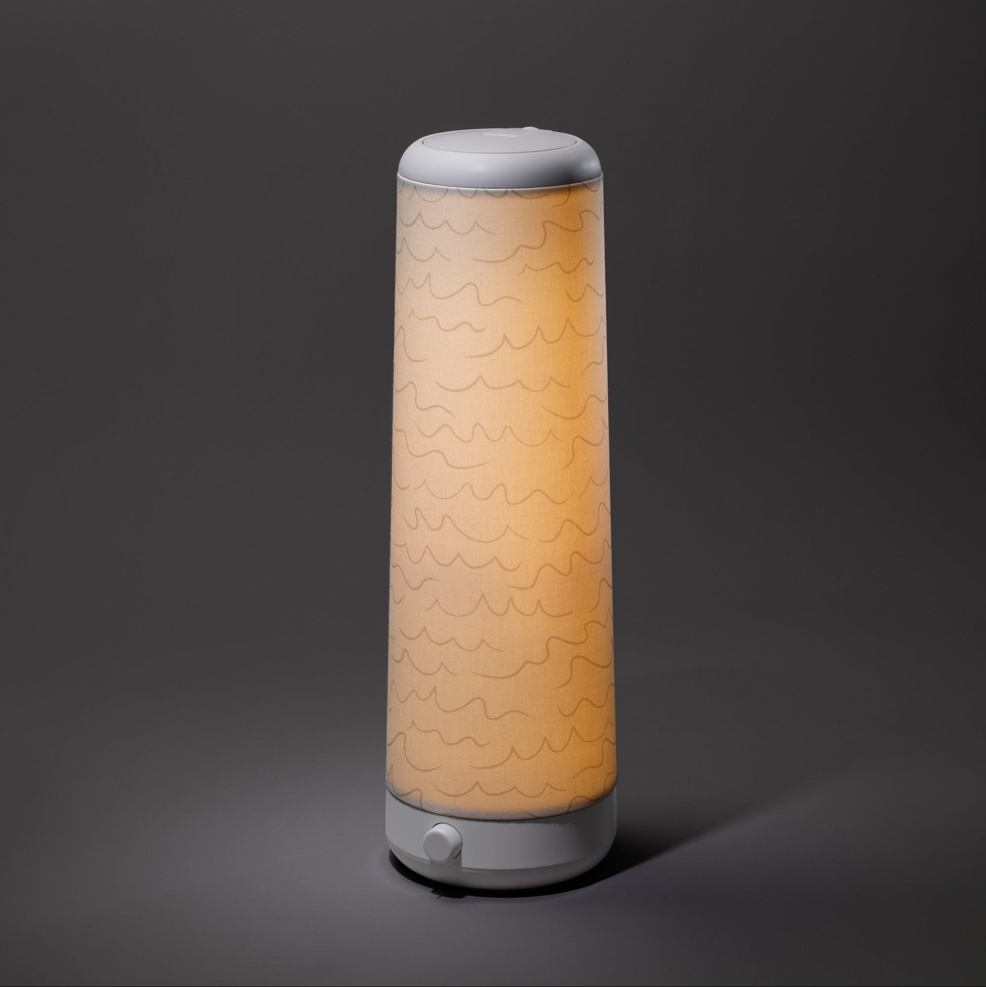 The Nightside Cursive Lines Cotton Shade lamp features a modern cylindrical design, a white base and top, interchangeable fabric shades with subtle wavy lines, and emits a soft, warm light against a dark background.