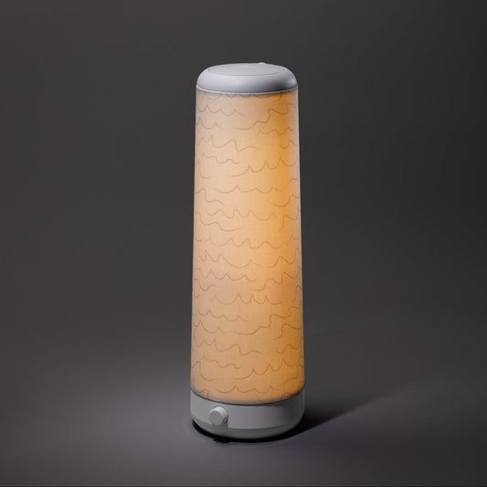 The Nightside Cursive Lines Cotton Shade lamp features a modern cylindrical design, a white base and top, interchangeable fabric shades with subtle wavy lines, and emits a soft, warm light against a dark background.