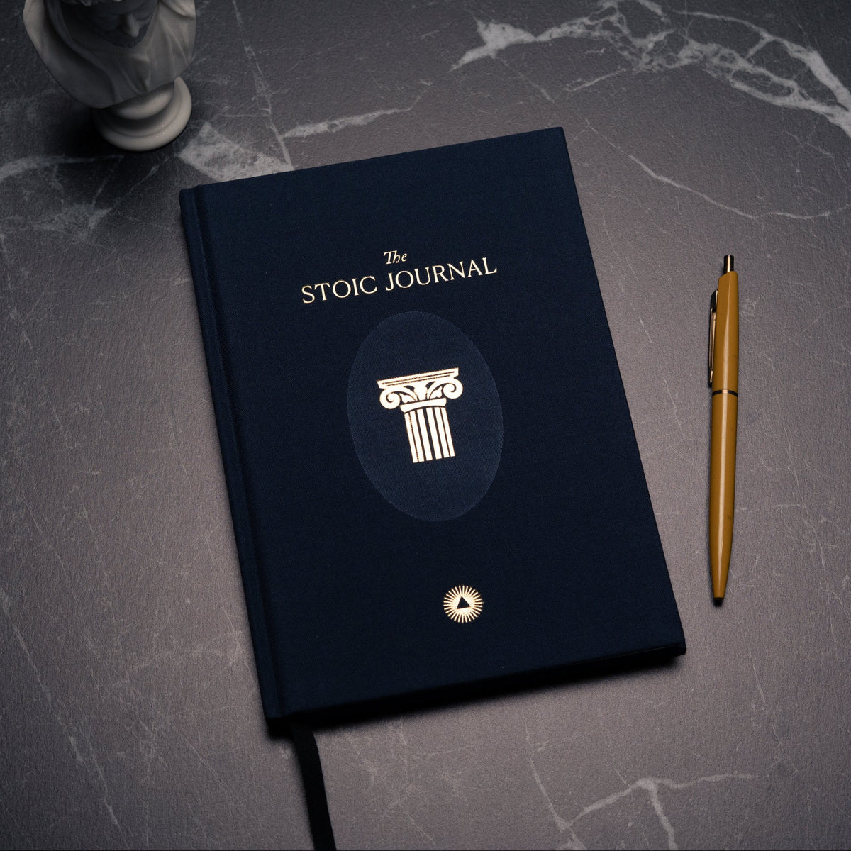 A dark navy blue Stoic Journal with gold lettering and a column emblem, shown closed on a dark marble surface next to a gold pen.