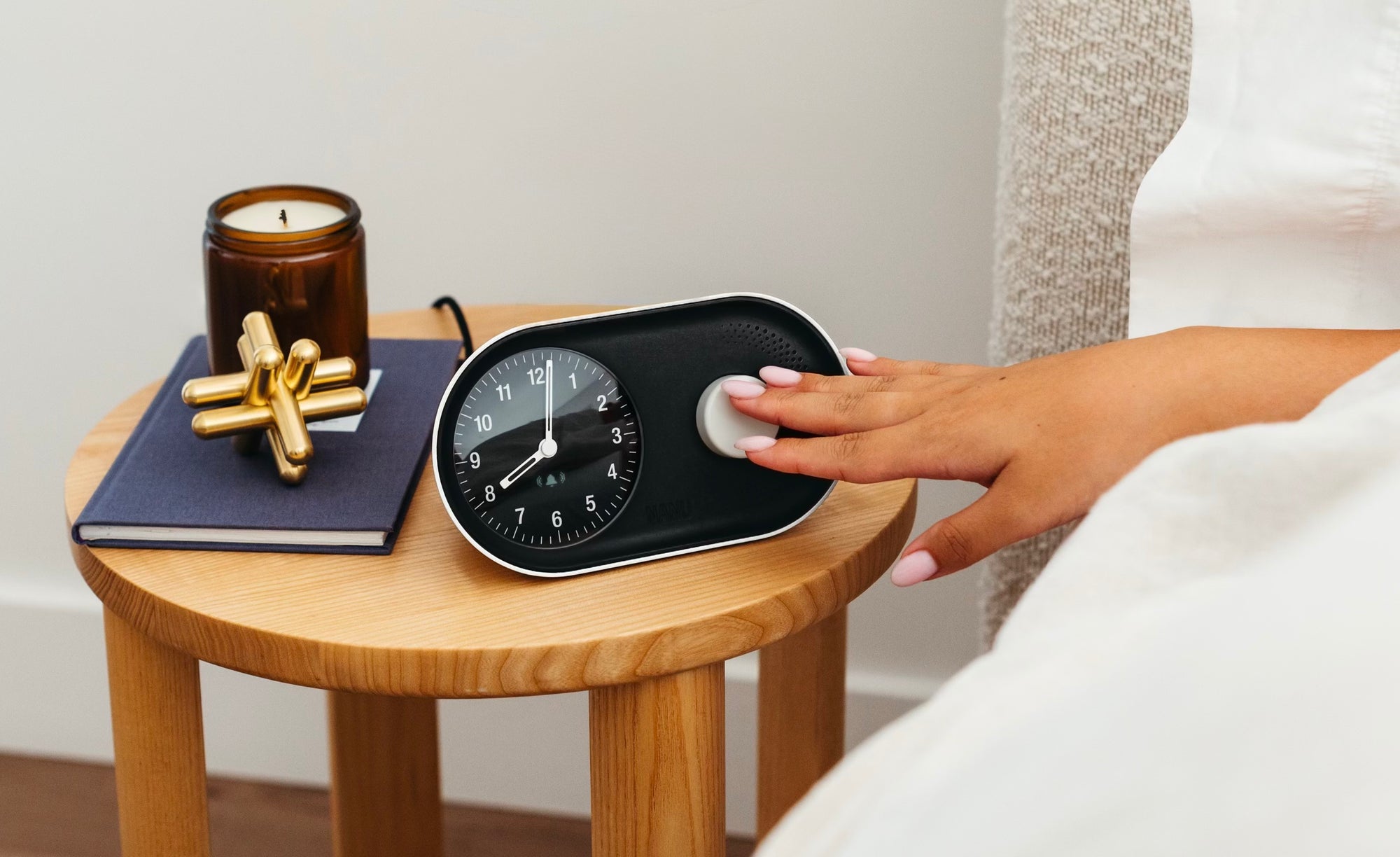 A persons hand gently reaches to turn off the Nanu Arc Alarm Clock on a wooden bedside table. The table also features a closed book, a gold decorative piece, and a lit candle in a brown glass holder.