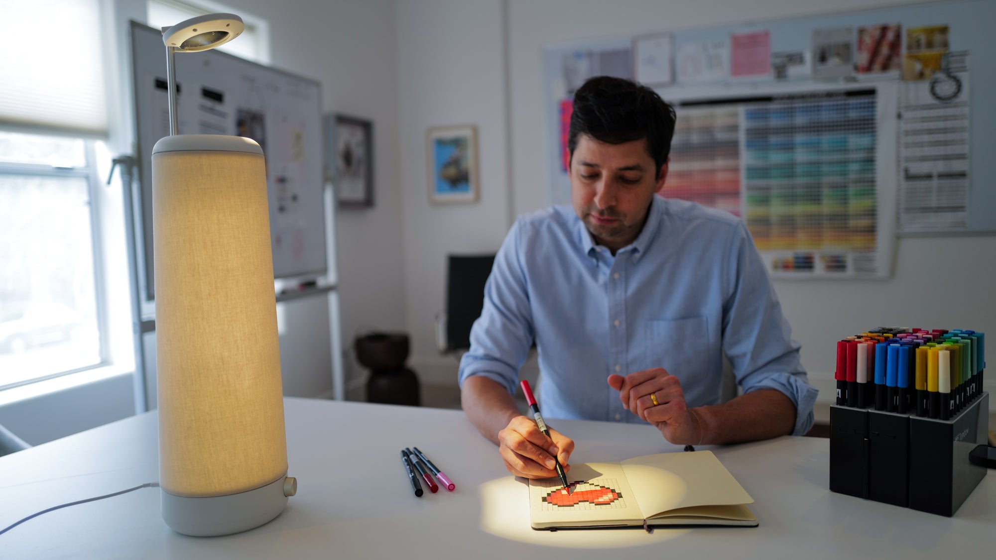 A person in a light blue shirt is seated at a desk, drawing in a notebook under a desk lamp. The desk has several colored pens, and the background features a whiteboard and a color chart on the wall.