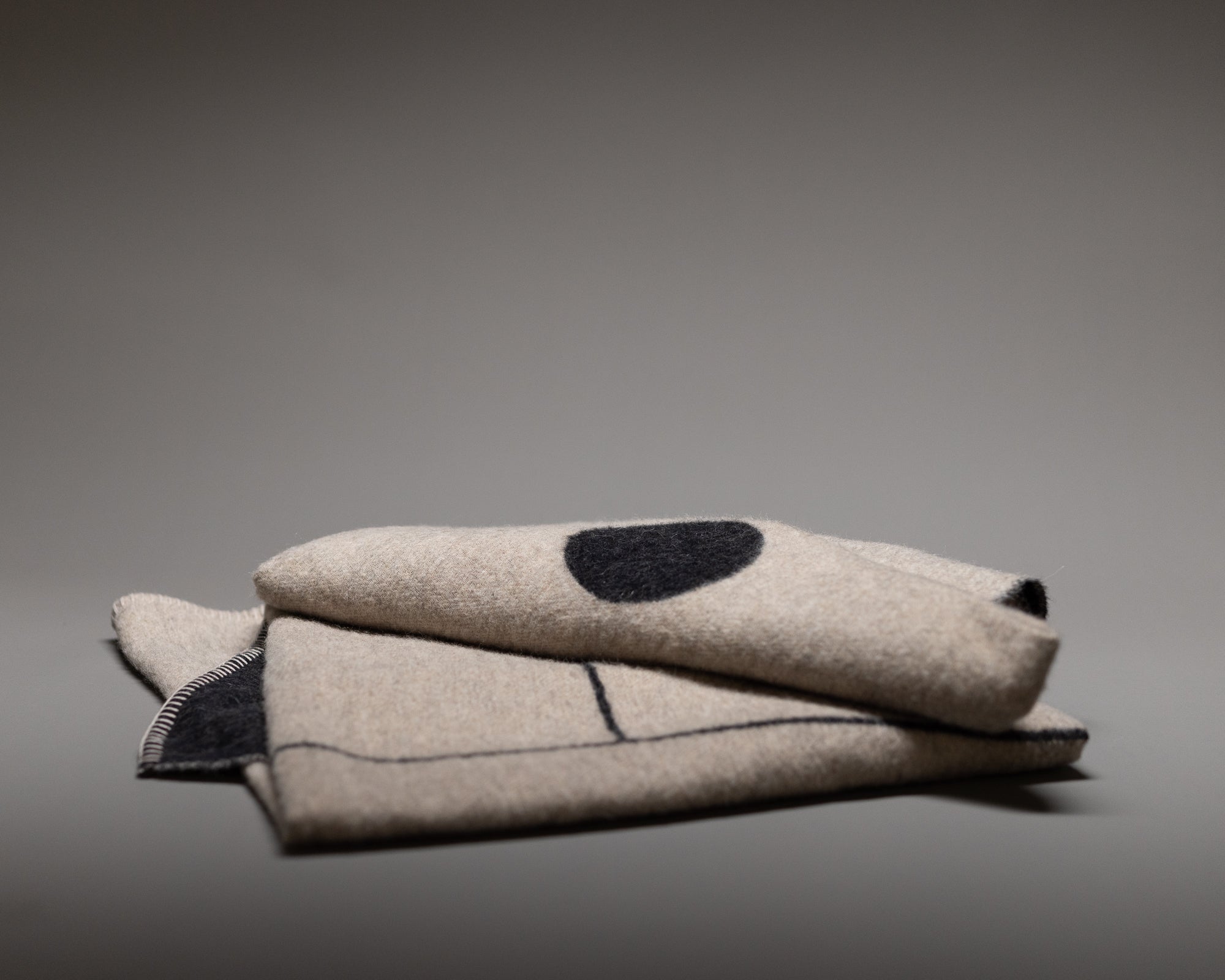 The Blacksaw Midnight Sun Throw Blanket is a folded beige wool piece with black stitched lines and a large black dot, made from eco-friendly sustainable fibers, shown on a plain gray background.