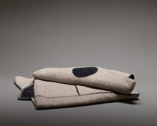The Blacksaw Midnight Sun Throw Blanket is a folded beige wool piece with black stitched lines and a large black dot, made from eco-friendly sustainable fibers, shown on a plain gray background.