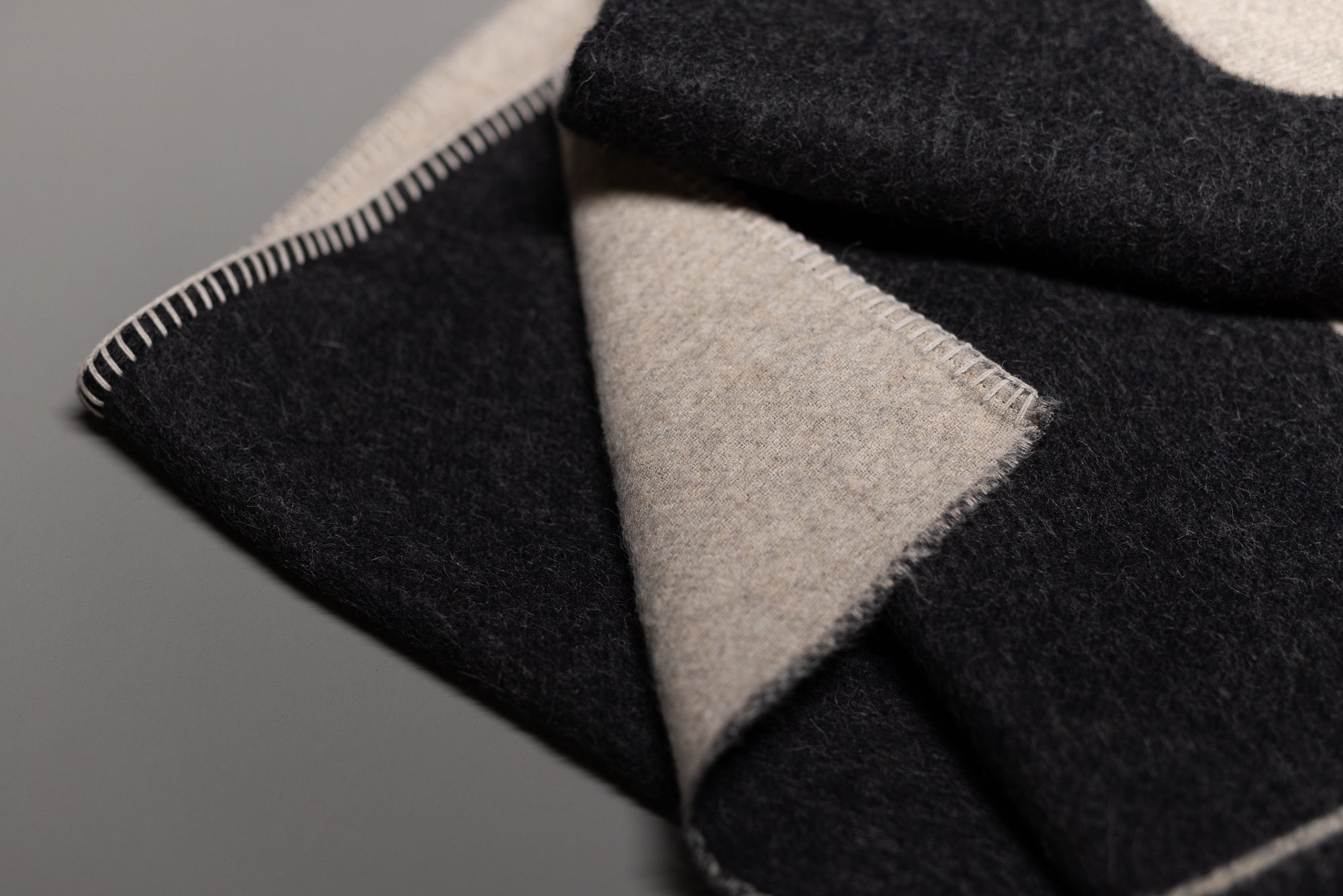 A close-up of the Blacksaw Midnight Sun Throw Blanket, featuring a folded, hypoallergenic wool design with black and beige hues, detailed edge stitching, and soft texture on both sides, set against a plain background.