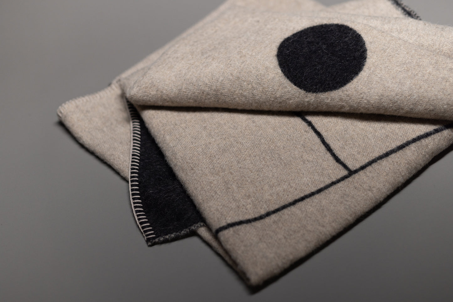 A close-up of the folded Midnight Sun Throw Blanket by Blacksaw, crafted from eco-friendly wool with black stitched edges and a minimal geometric pattern, shown on a smooth gray surface.