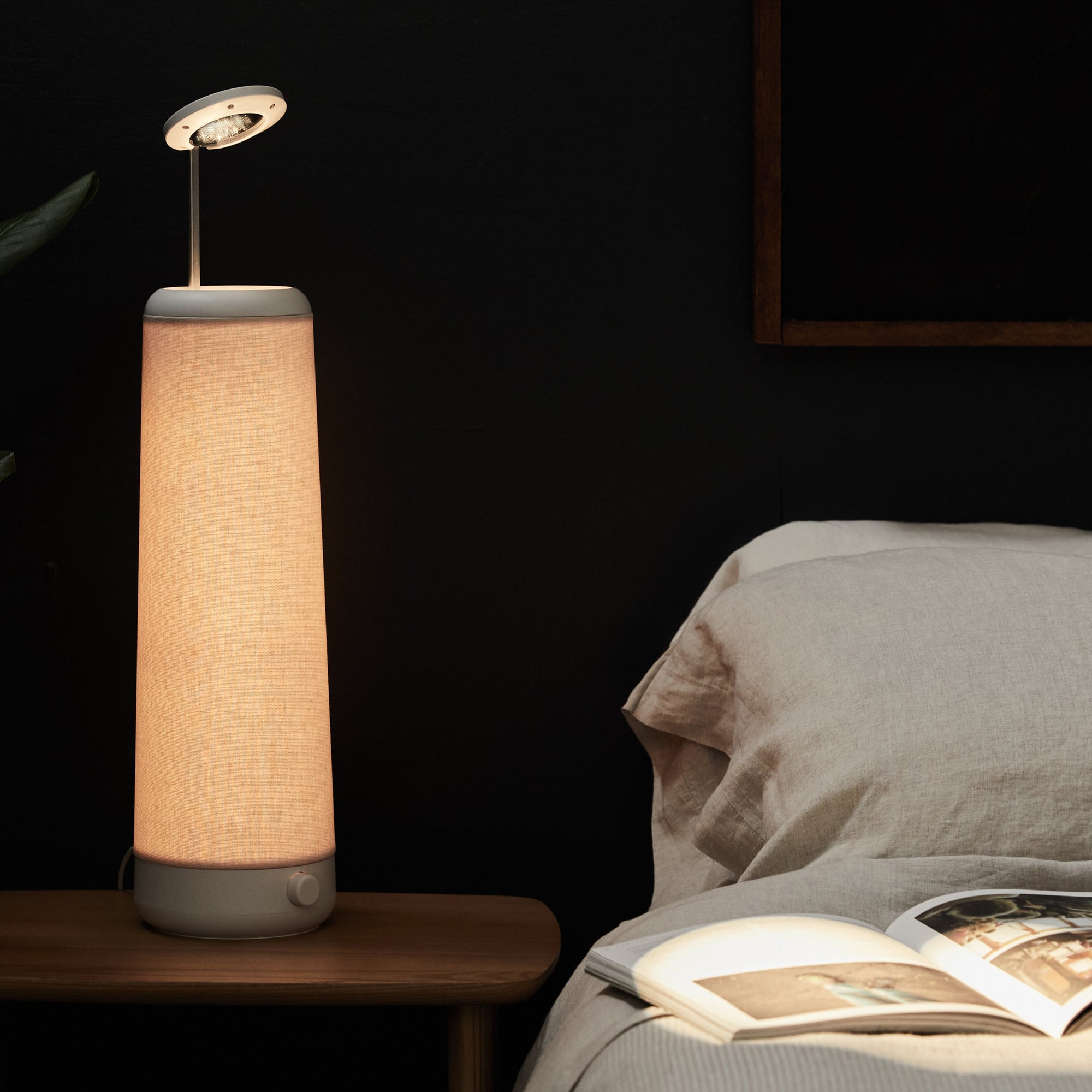 A tall, cylindrical Nightside NS01 Personal Task Lamp with a soothing Warm-White LED and a light brown shade sits on a wooden bedside table. Its gentle glow contrasts with the dark background wall, illuminating an open book on the bed beside a beige pillow.