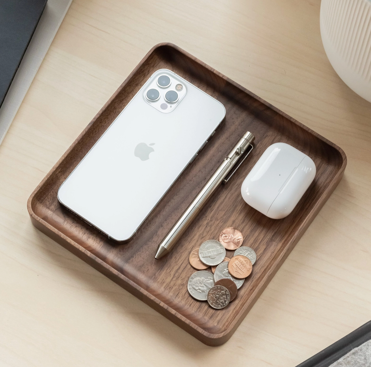 The Ugmonk Valet Tray - Medium, crafted from precision-milled walnut, sits on a light desk holding a silver iPhone, metallic pen, white wireless earbuds in a case, and assorted coins—showcasing its unique grain and elegant design.