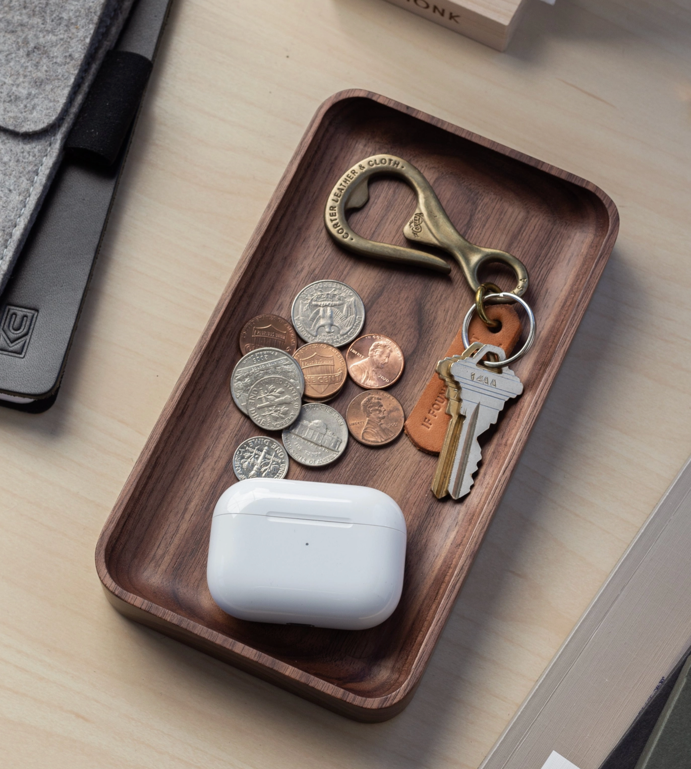 On the desk, the Ugmonk Valet Tray - Small, crafted from solid walnut, organizes coins, a brass bottle opener, two keys on a leather keychain, and a white wireless earbuds case. A gray sleeve and other items are nearby.