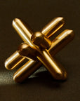 The Brass Jack Puzzle by Craighill is a gold metallic sculpture crafted from solid brass, resembling interlocking rods. Measuring 3x3x3 with a 1/2 diameter, it sits on a dark textured surface, reflecting light off its smooth, shiny finish and highlighting its three-dimensional shape.